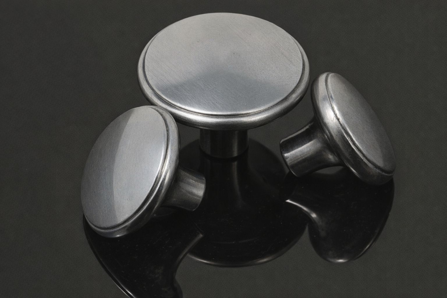 Kolme pyöreää harjattua teräsnuppia mustalla heijastavalla alustalla.